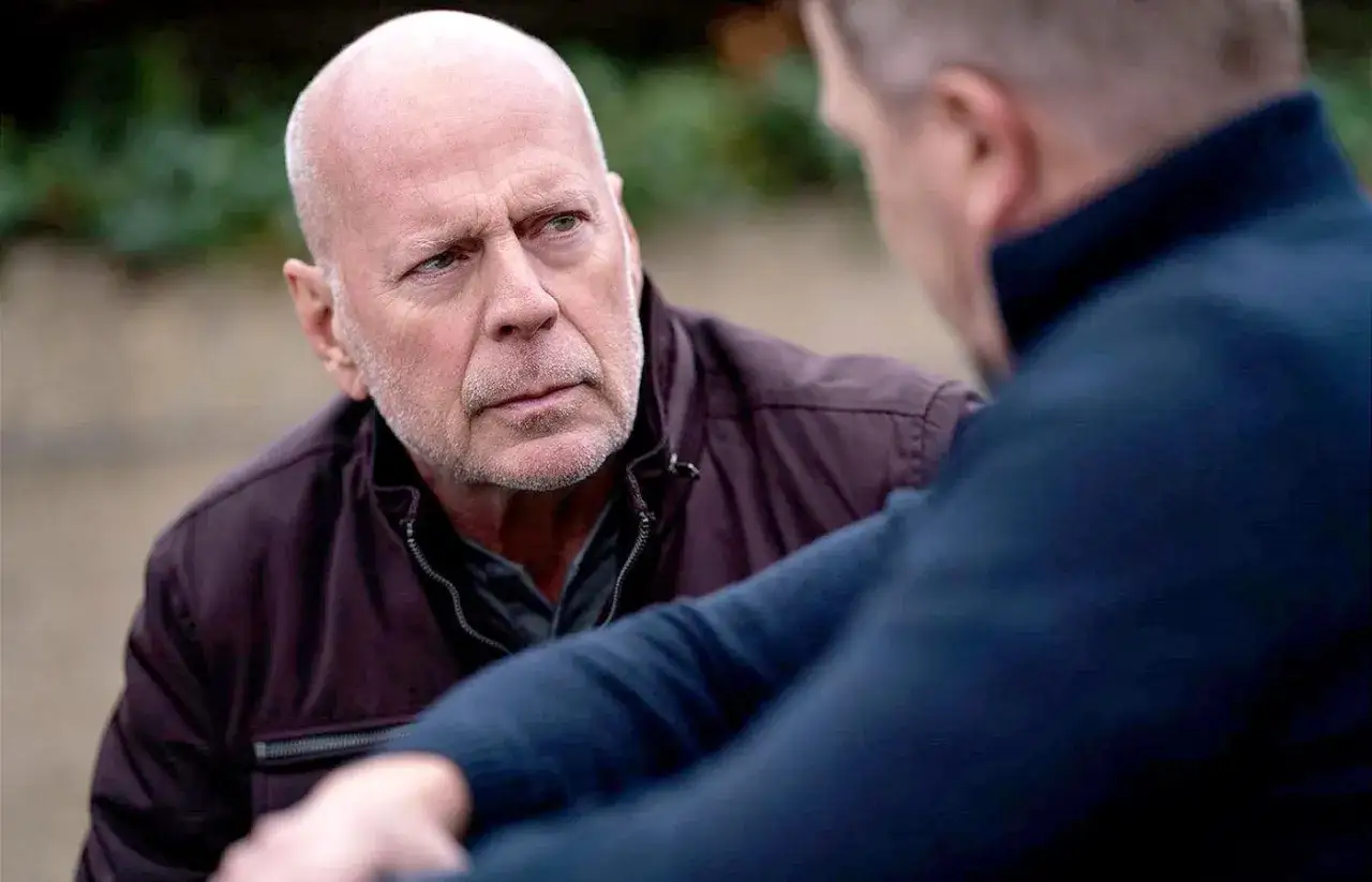 Bruce Willis w rozmowie, jego twarz wyraża skupienie. W tle widać fragment innej osoby. To trudny czas dla aktora, gdy zmaga się z chorobą.