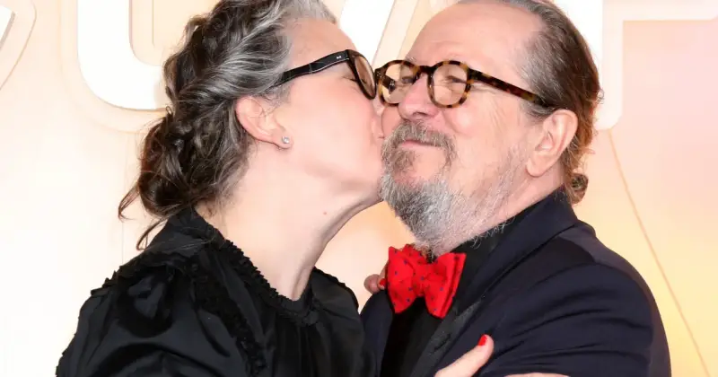 Gary Oldman otrzymuje czuły pocałunek od swojej żony. Oboje mają okulary, a ona nosi czarną sukienkę.